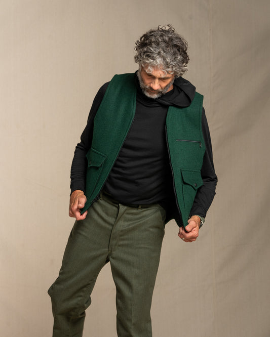 Man wearing a green wool vest over a black merino wool shirt and green pants against a beige background