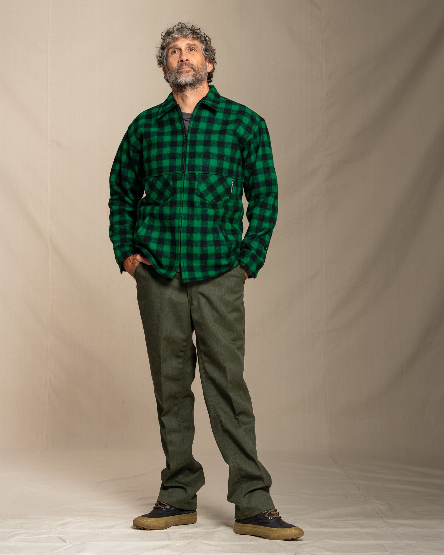 Man wearing a green and black checkered wool shirt and green pants against a beige background