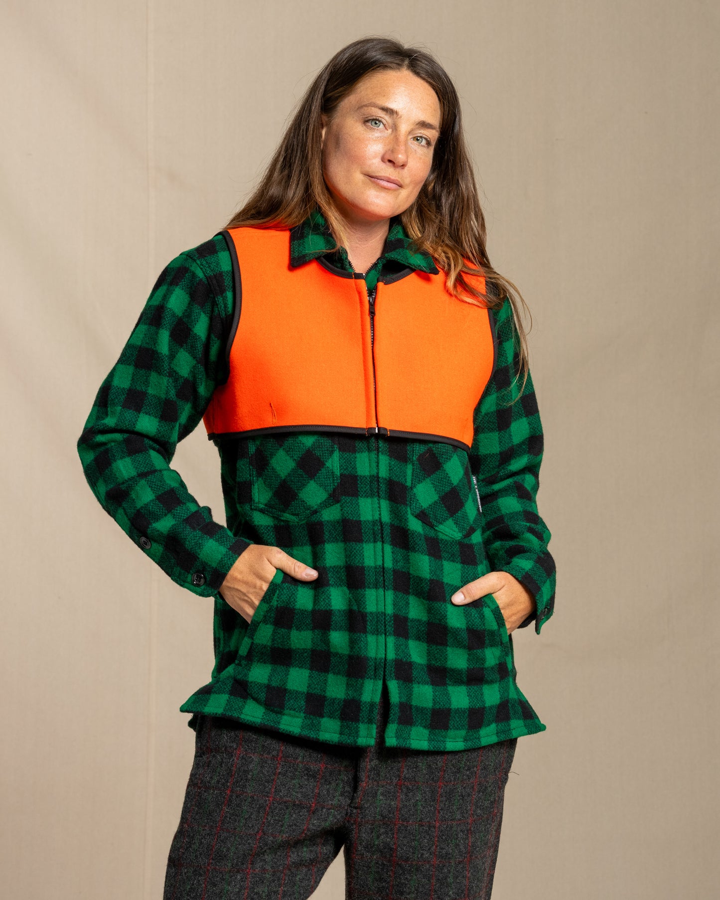 Person wearing a green and black checkered jacket with an orange wool safety vest on a beige background