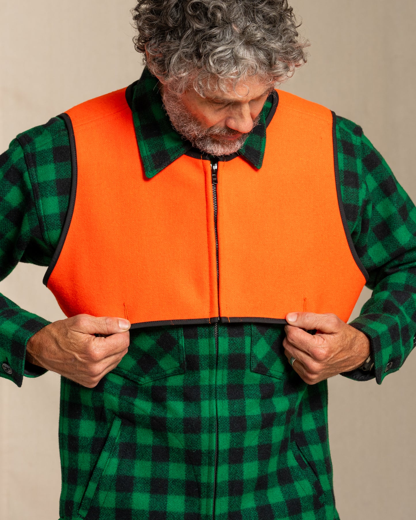 Man wearing a green and black plaid shirt with an blaze orange wool safety vest over it on a beige background