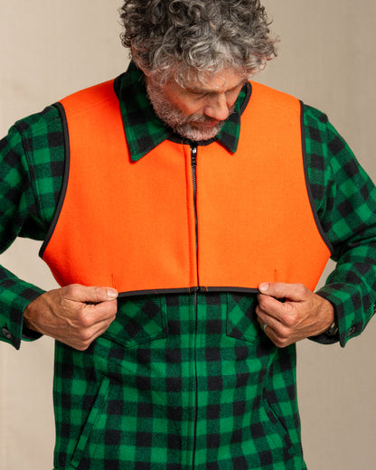 Man wearing a green and black plaid shirt with an blaze orange wool safety vest over it on a beige background