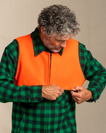 Man wearing a green checkered shirt and blaze orange wool safety cape against a beige background
