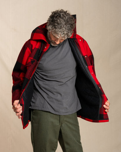 Person wearing a red and black checkered jacket over a gray shirt and green pants on a beige background