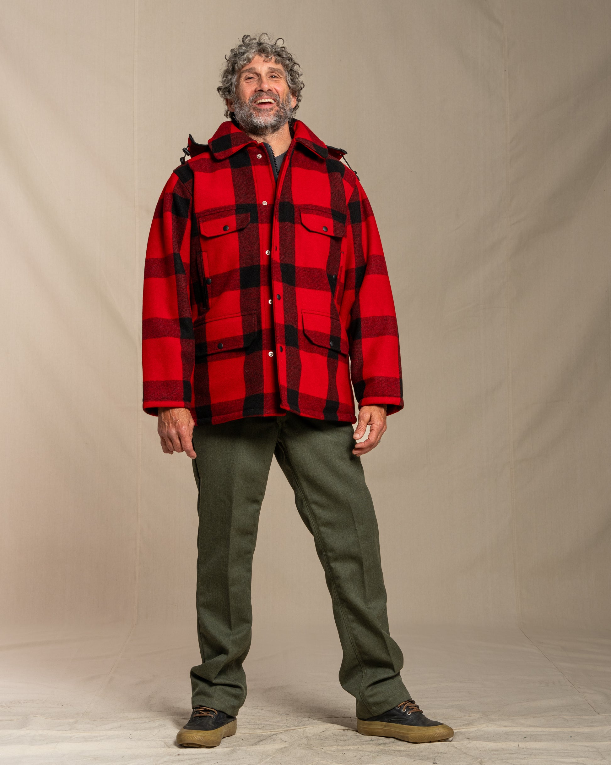 Man wearing a red and black plaid mackinaw wool jacket over a black shirt, standing against a plain background