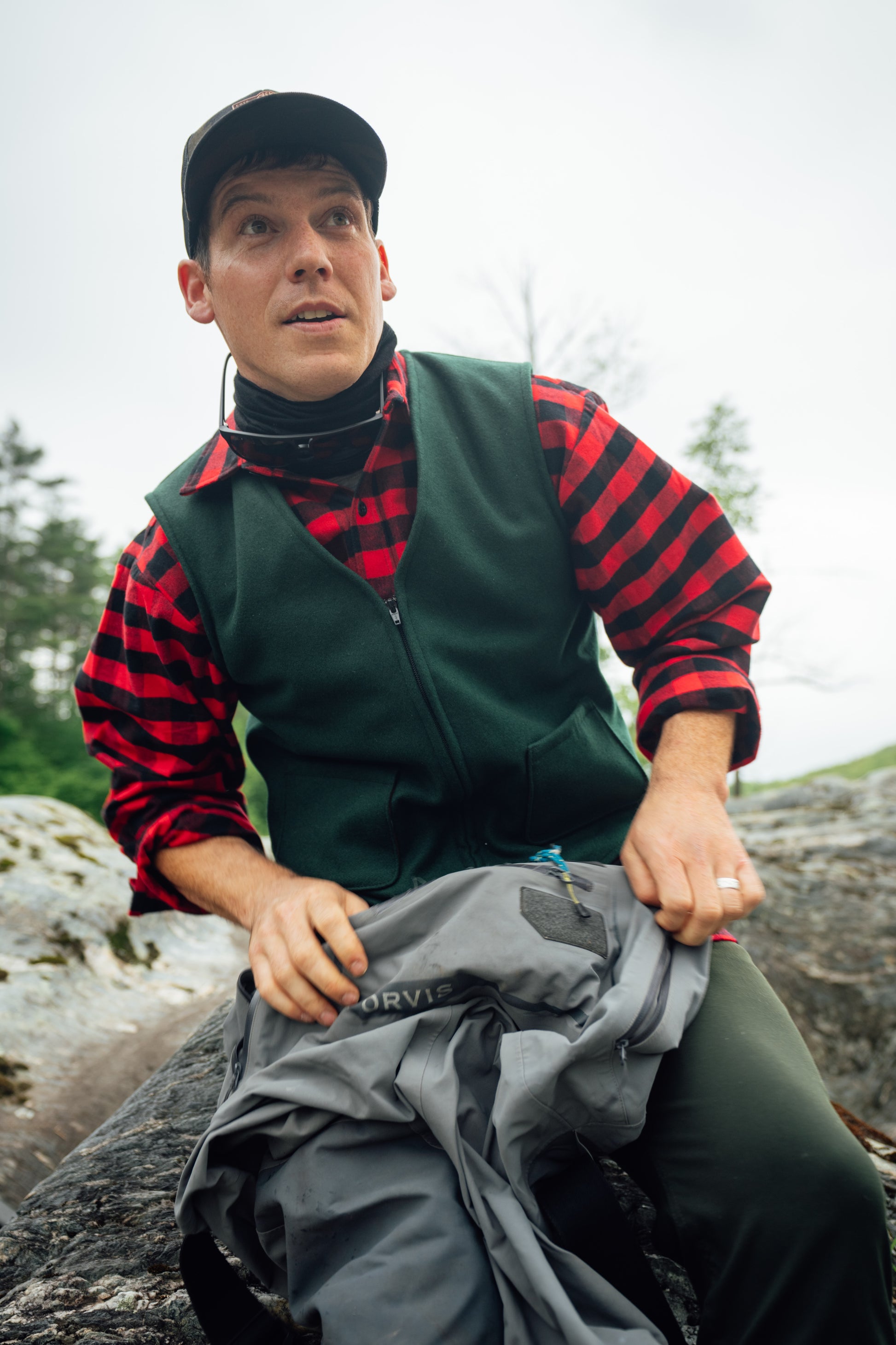 A man wearing a green vest over a red and black striped flannel shirt sits outdoors on a rock, holding a gray Marvis jacket.
