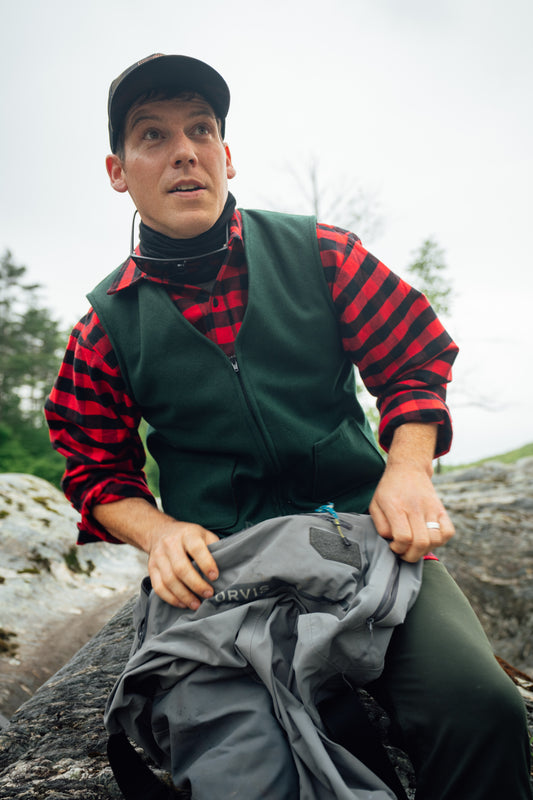 A man wearing a green vest over a red and black striped flannel shirt sits outdoors on a rock, holding a gray Marvis jacket.