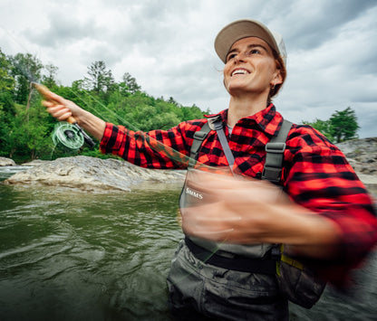 A woman fly fishing in a river, wearing a red and black plaid shirt, gray waders, and a beige cap, while casting her line. The background shows a rocky river and a green forest under a cloudy sky.