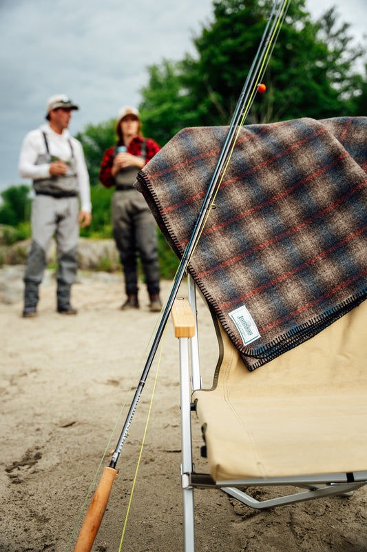 Plaid brown, blue, red blanket over beach chair on beach with fly rod leaning against arm rest. Couple in the background with waiters taking in a drink after fishing
