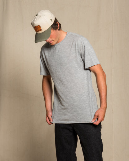 A man wearing a light gray heather crewneck merino wool t-shirt, black pants, and a cream and gray baseball cap, standing against a neutral beige wall.