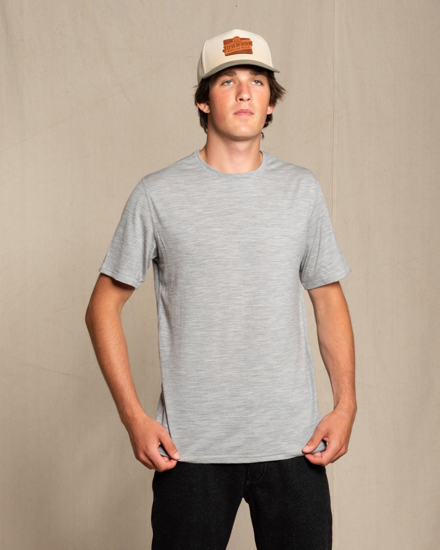 A man wearing a light gray heather crewneck merino wool t-shirt and a beige baseball cap with a brown logo patch, standing against a neutral beige backdrop.