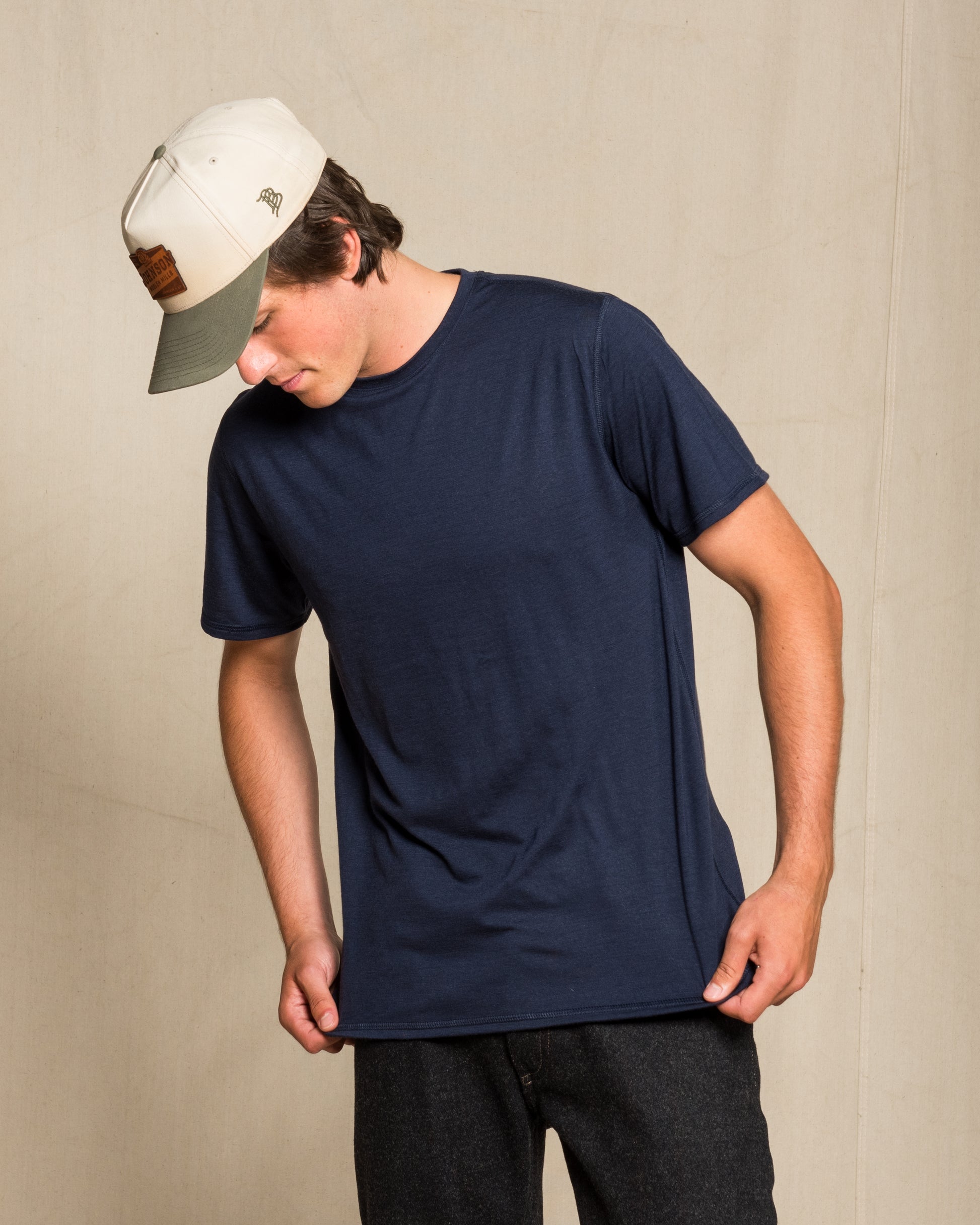 A man wearing a navy blue crewneck merino wool t-shirt, dark gray pants, and a white baseball cap with a brown patch, standing against a neutral beige background.