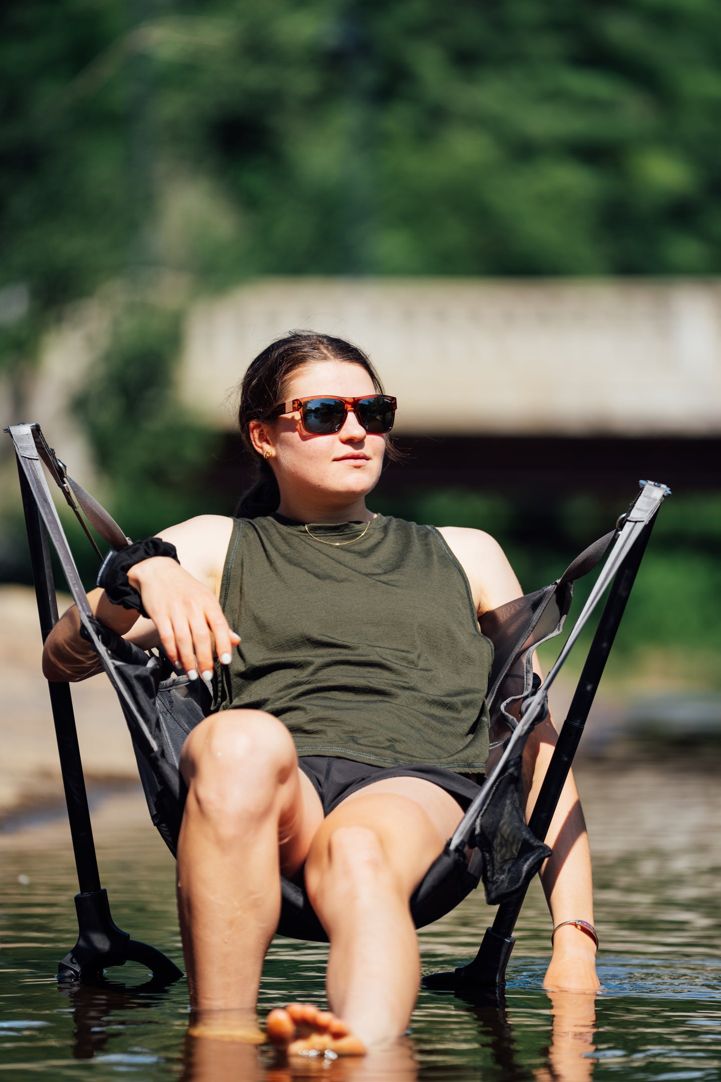 A woman wearing sunglasses and an olive green tank top sits in a black mesh reclining chair with armrests, with her legs dangling into shallow water outdoors.