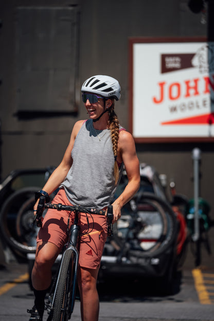 A woman riding a bicycle outdoors, wearing a white helmet, black sunglasses, a gray merino wool sleeveless top, and rust-orange shorts with black graphics. A large sign reading 'Johnson Woolen Mills'.