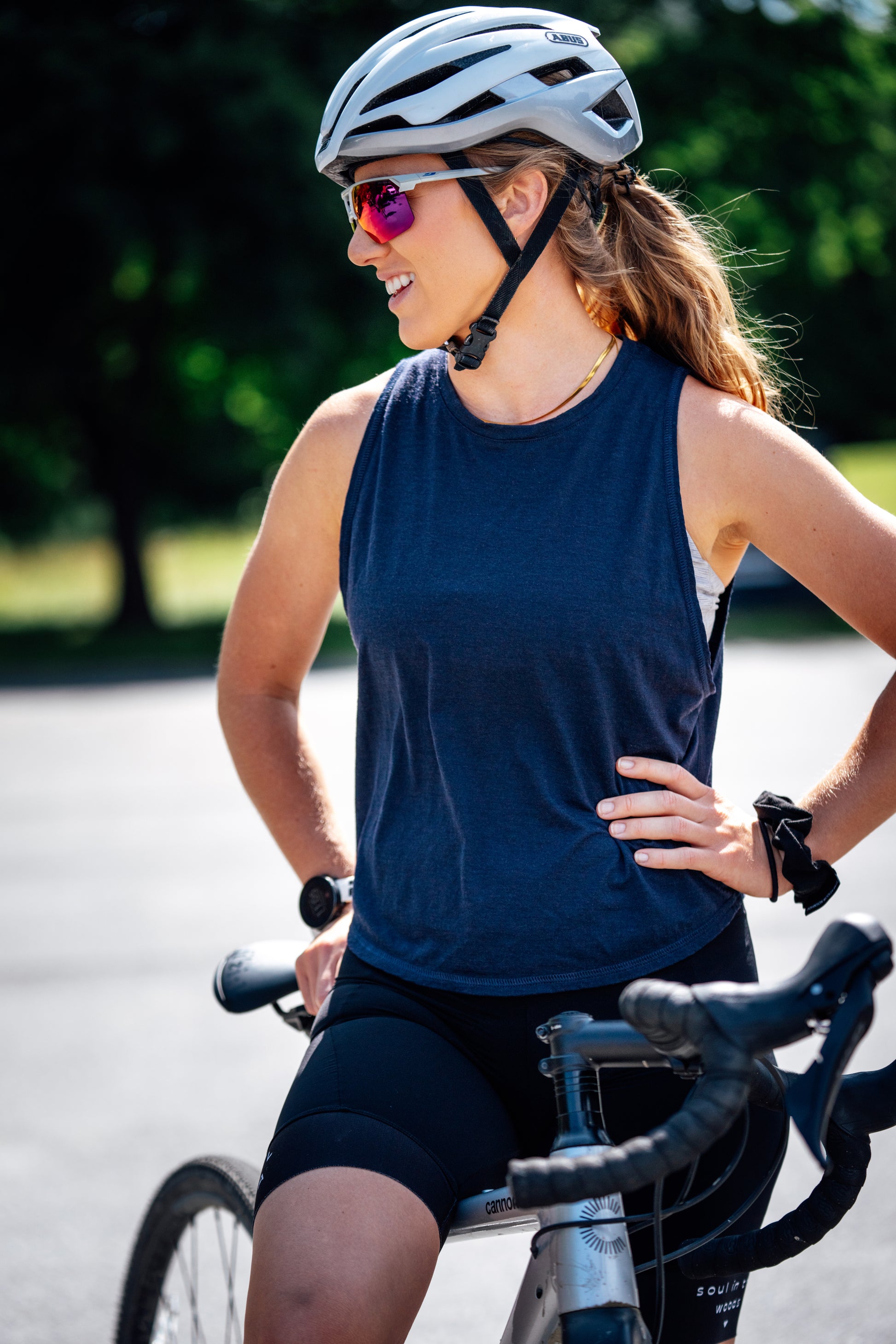 A woman wearing a white bicycle helmet, pink-tinted sunglasses, a navy blue merino wool sleeveless cycling tank top, and black cycling shorts, posing on a road bike outdoors.