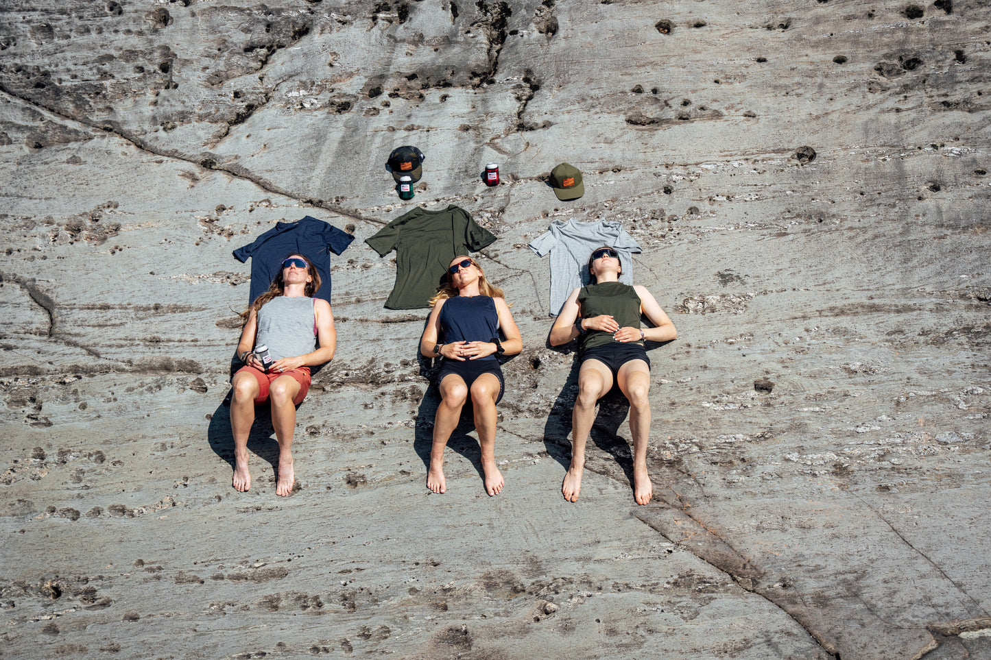 Three women are lying on a large rock face wearing merino wool tank tops, with several t-shirts and hats laid out above them on the rock. The photo is taken from a high-angle overhead view.