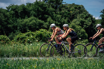 Three cyclists riding gravel bikes on a grassy path with a dense green forest in the background under a blue sky.