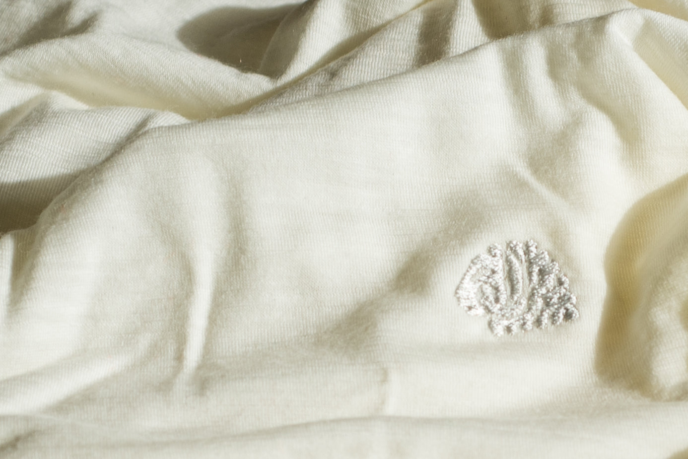 A close-up of cream-colored merino wool fabric with a small white embroidered monogram logo on the right side.