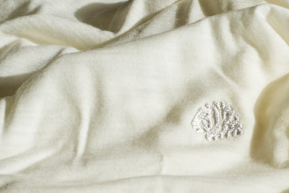 A close-up of cream-colored merino wool fabric with a small white embroidered monogram logo on the right side.