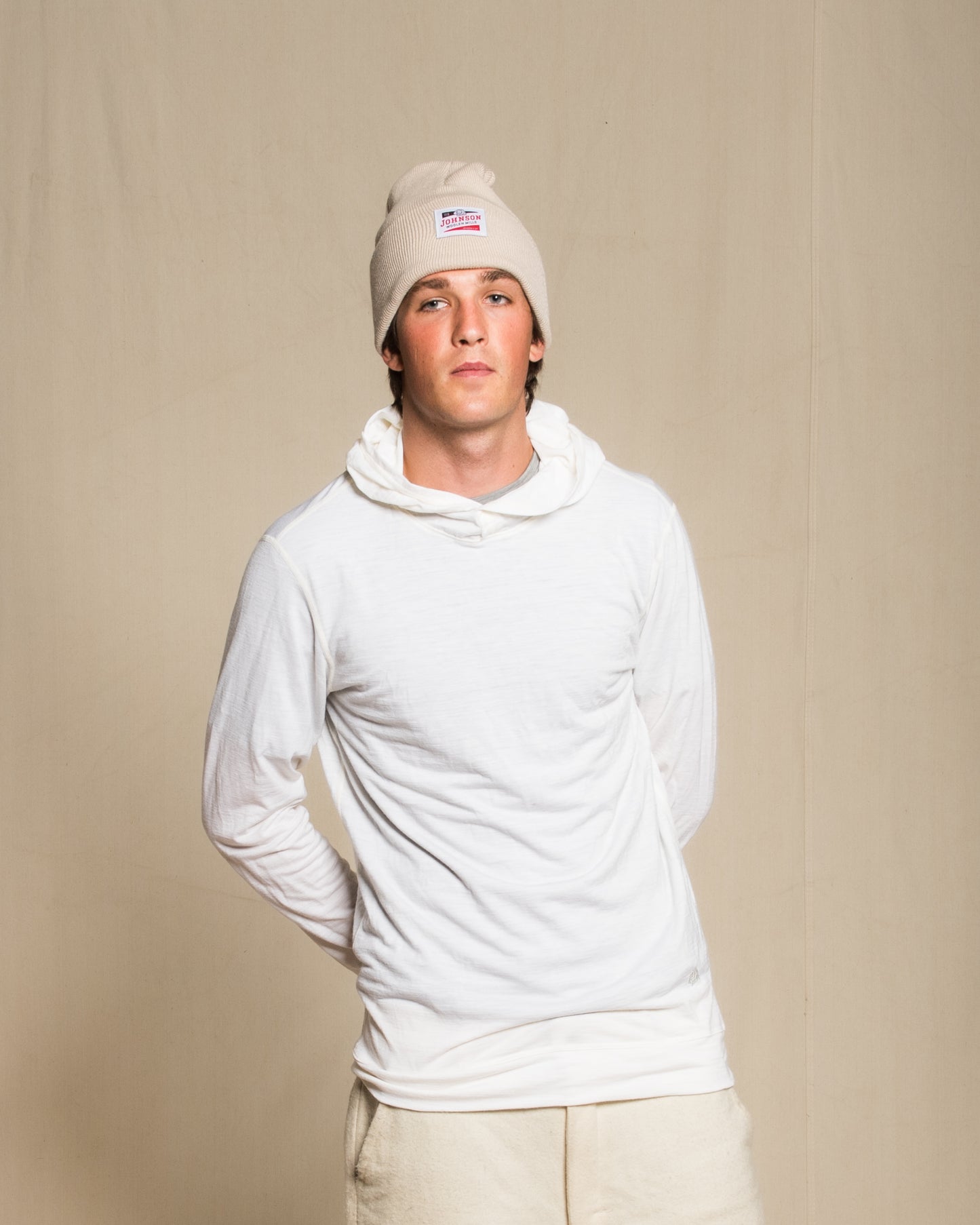 A man wearing a light beige knit beanie, a white merino wool long-sleeve hooded shirt, and light beige pants, standing against a neutral beige backdrop.