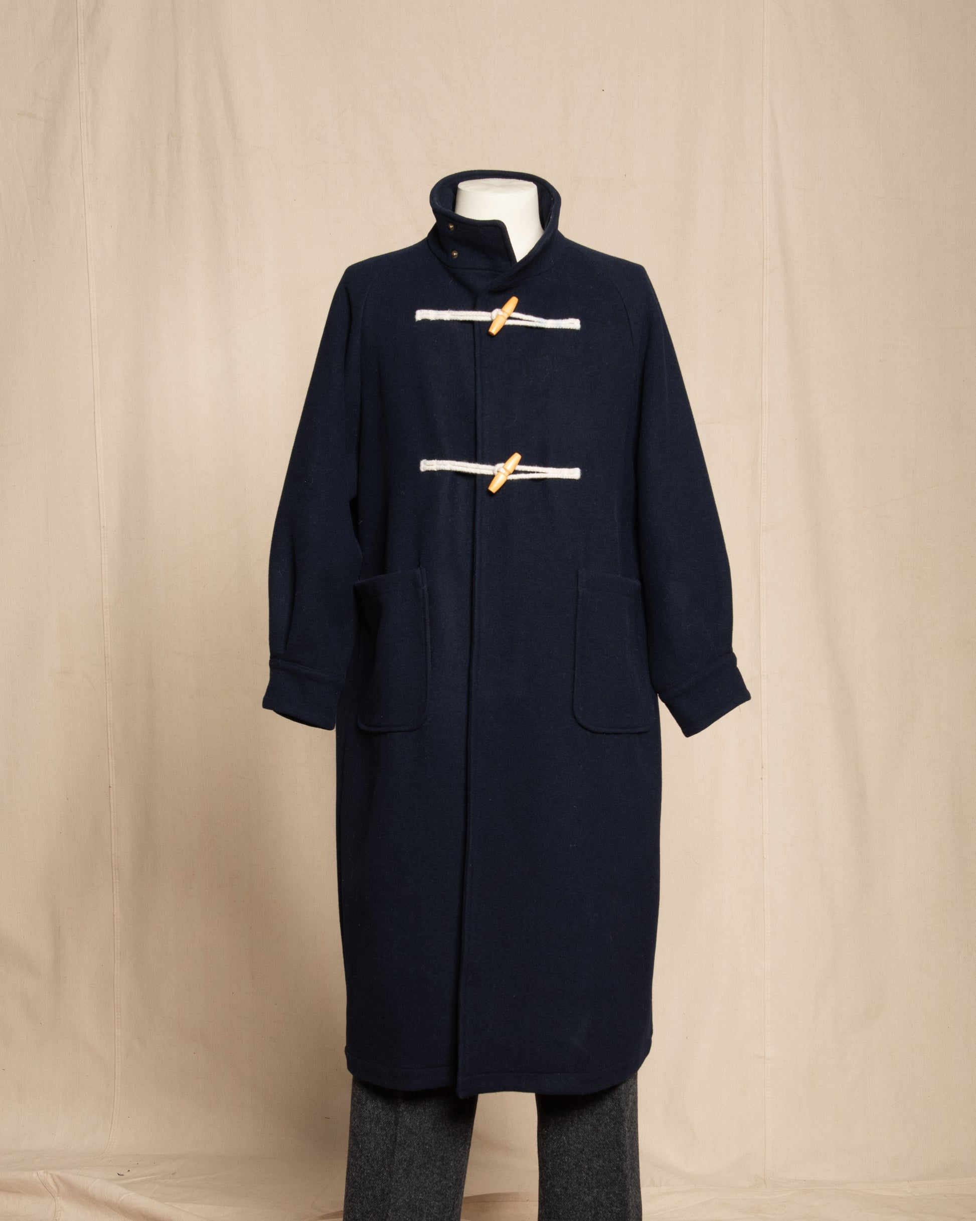 A dark navy blue wool duffle coat with a high collar, displayed on a mannequin against a beige fabric backdrop. The coat features two front pockets and is fastened with two horizontal white rope toggles tied with tan leather loops.