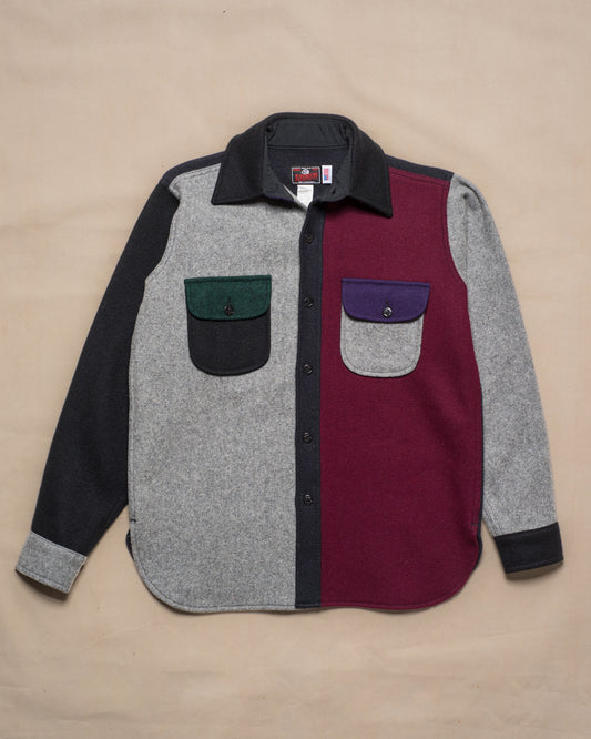 A color-blocked long-sleeve wool button-up shirt laid flat on a beige background. The shirt features a heather gray body, a deep burgundy panel on the right side, black sleeves and collar, with a dark green chest pocket on the left and a purple chest pocket on the right.
