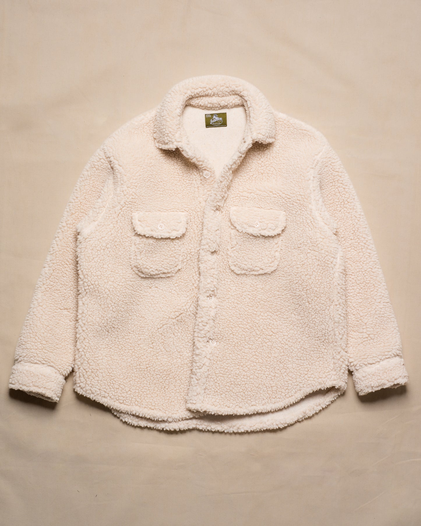 A cream-colored sherpa fleece button-up shirt jacket with two front flap pockets, laid flat on a beige fabric background.
