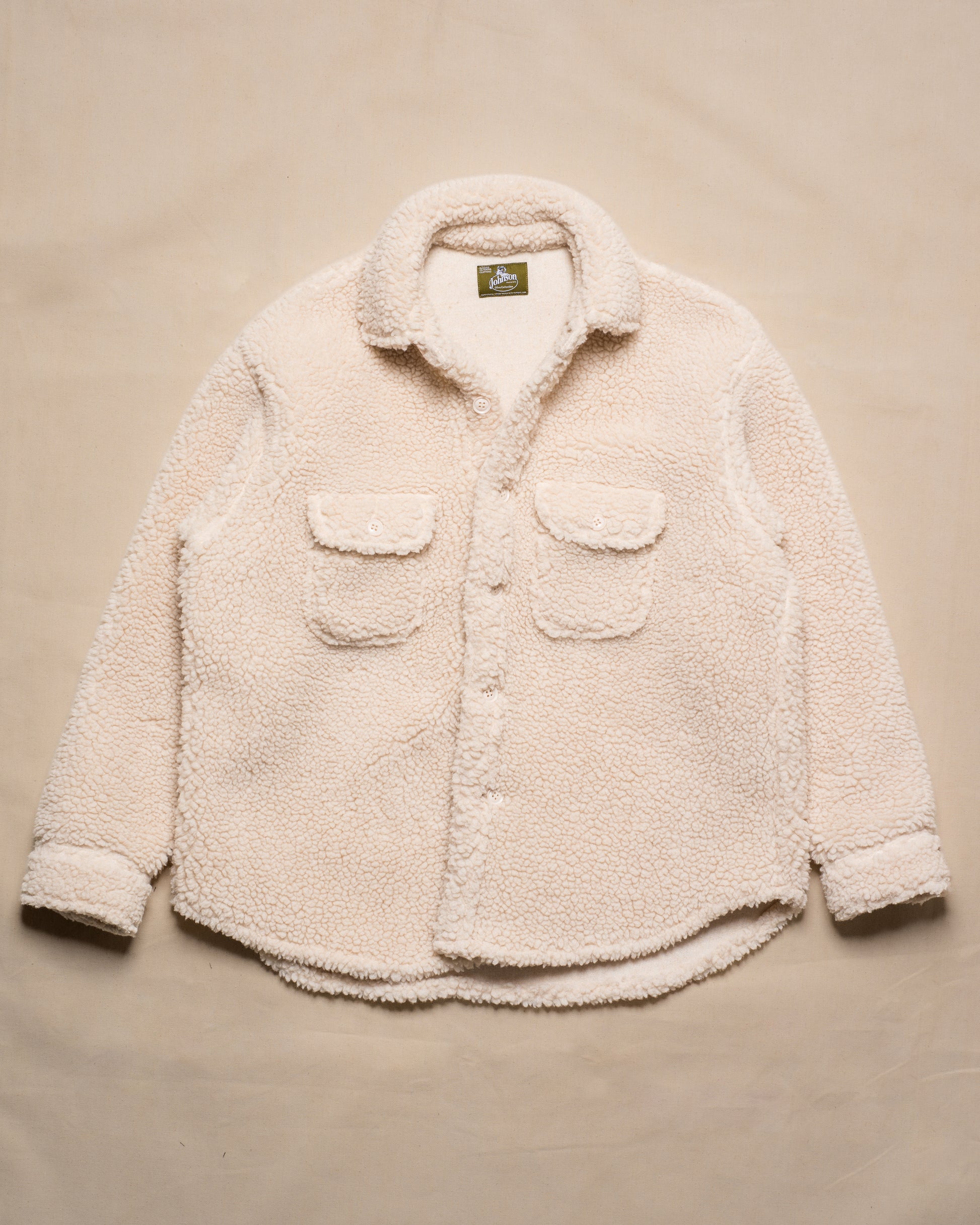 A cream-colored sherpa fleece button-up shirt jacket with two front flap pockets, laid flat on a beige fabric background.