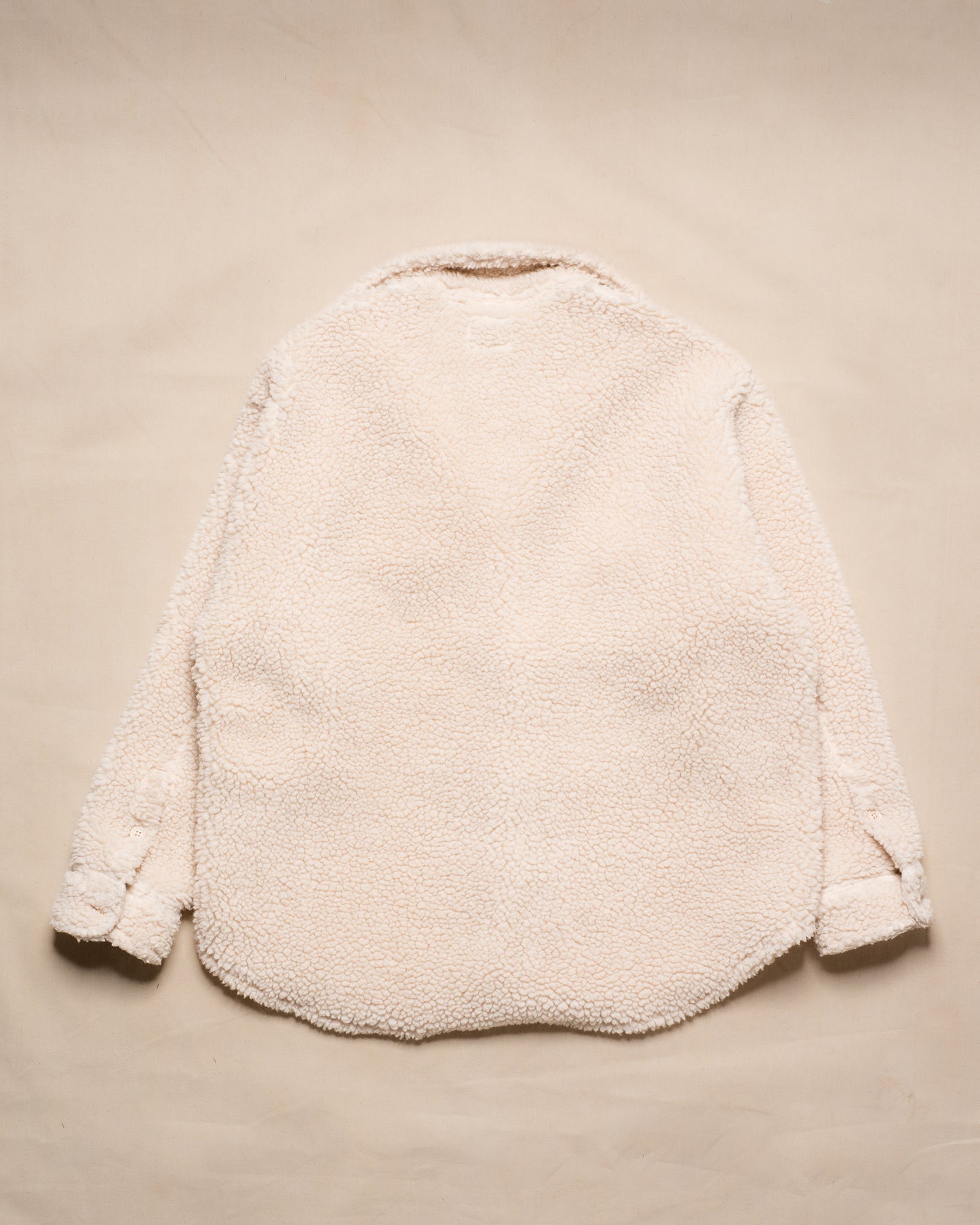 A cream-colored sherpa fleece jacket shown from the back, laid flat on a matching beige textured background.