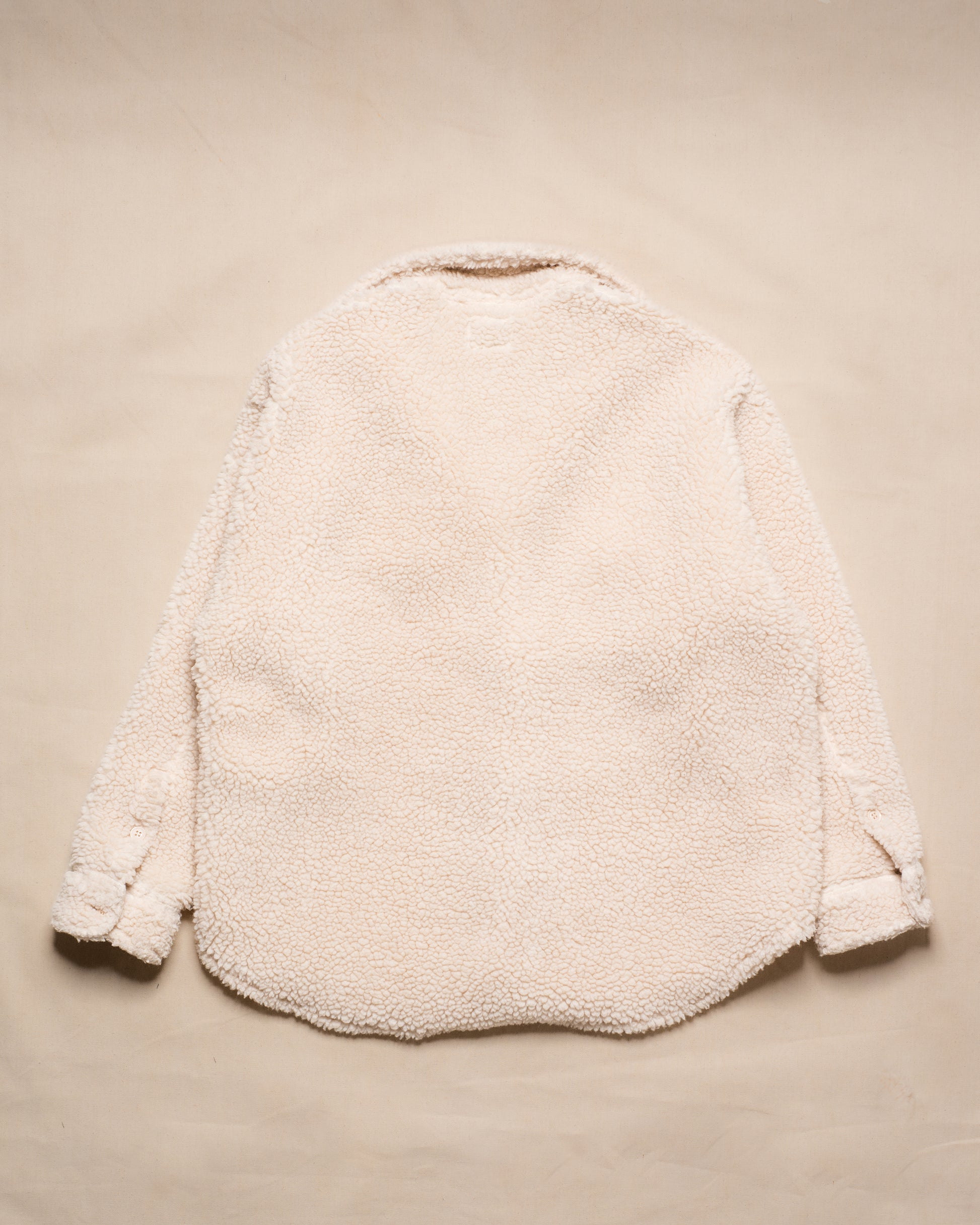 A cream-colored sherpa fleece jacket shown from the back, laid flat on a matching beige textured background.