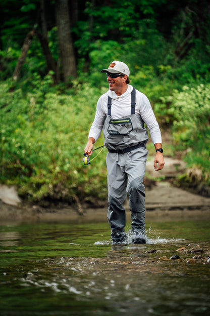 A man wearing gray fly-angling waders, a white merino wool hoodie, and a white cap stands in a shallow river, holding a fly fishing rod. The background is a lush green forest.