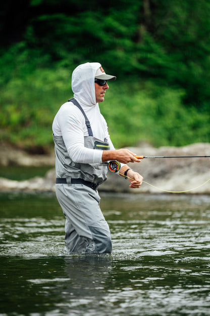 A man fly fishing in a river, wearing a white merino wool hoodie, gray waders, and a cap, with a blurred green forest background.