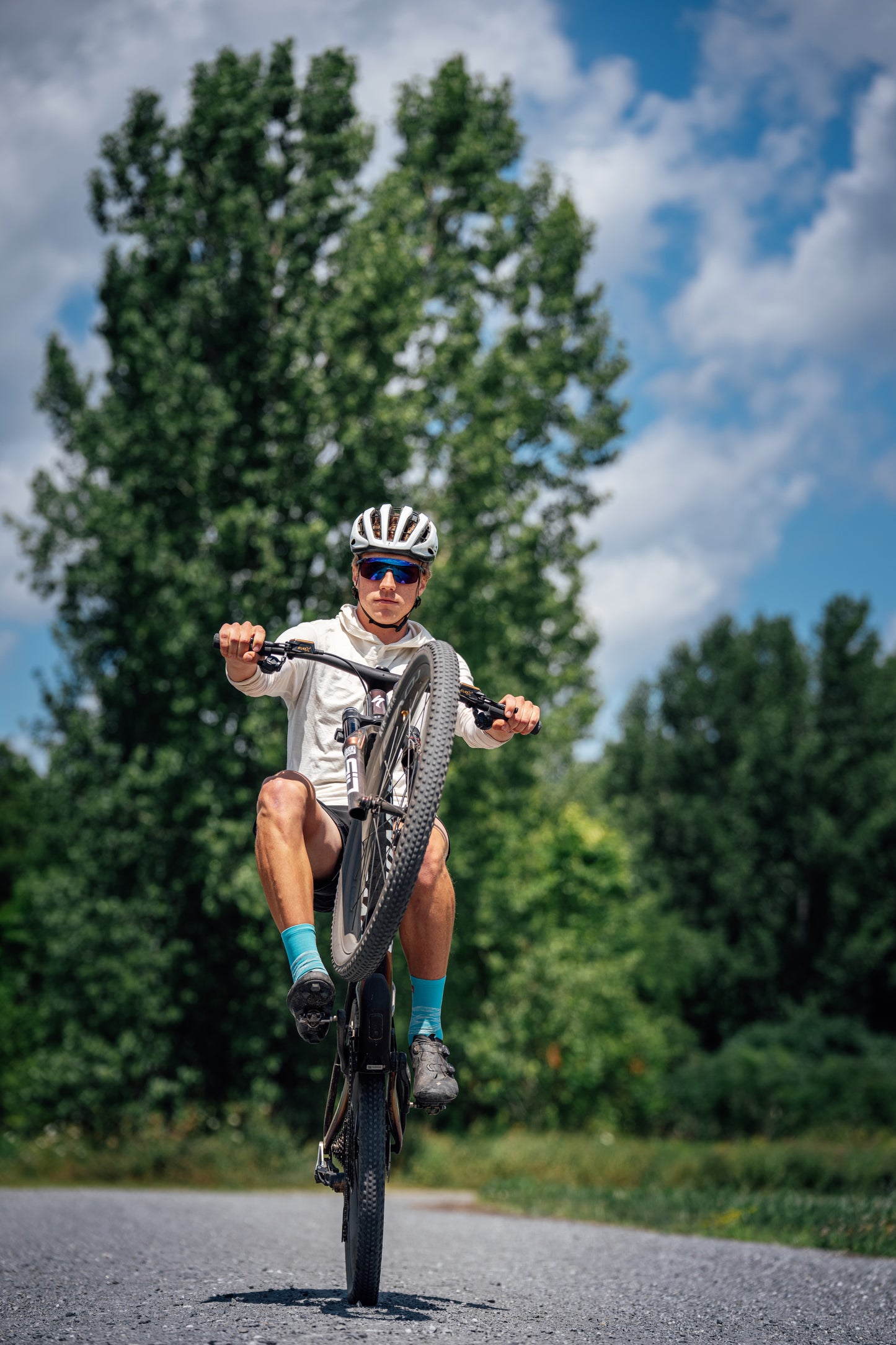 A man wearing a white helmet, sunglasses, and a white merino wool hoodie rides a mountain bike on a paved road outdoors with green trees and a blue sky in the background.