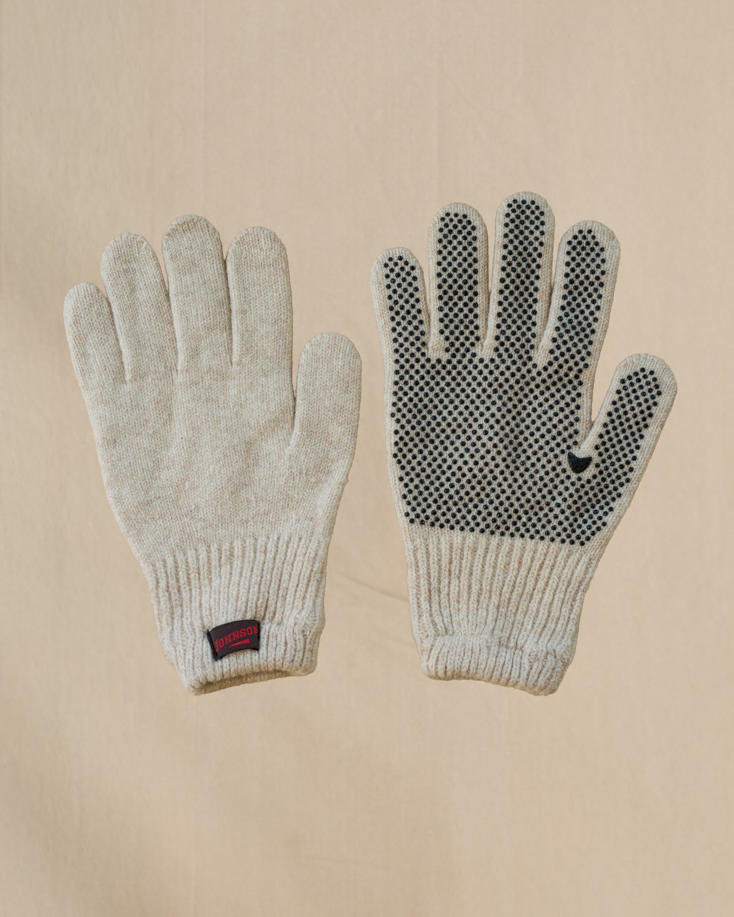 A pair of beige merino wool knit gloves shown flat against a light beige background, with one glove displaying a textured grip pattern on the back of the hand and a small brown and red Johnson tab on the cuff.