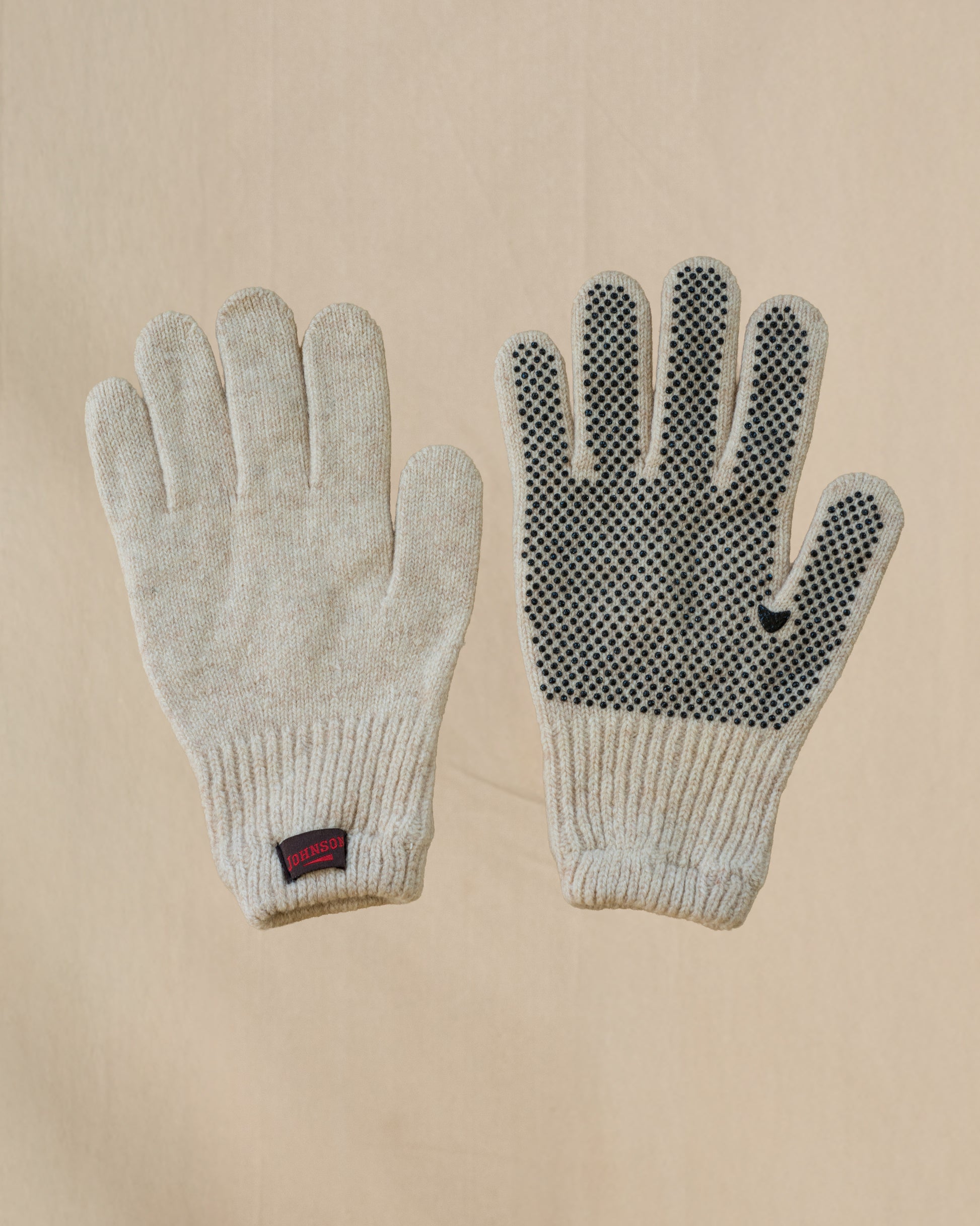 A pair of beige merino wool knit gloves shown flat against a light beige background, with one glove displaying a textured grip pattern on the back of the hand and a small brown and red Johnson tab on the cuff.