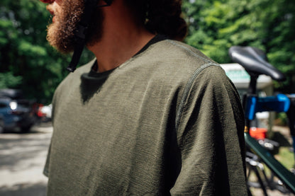 A close-up shot of a man wearing an olive crewneck merino wool shirt, standing outdoors with a bicycle visible in the background.
