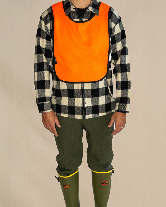 Person wearing a checkered shirt with an orange safety vest over it, green pants, and green boots on a beige background.