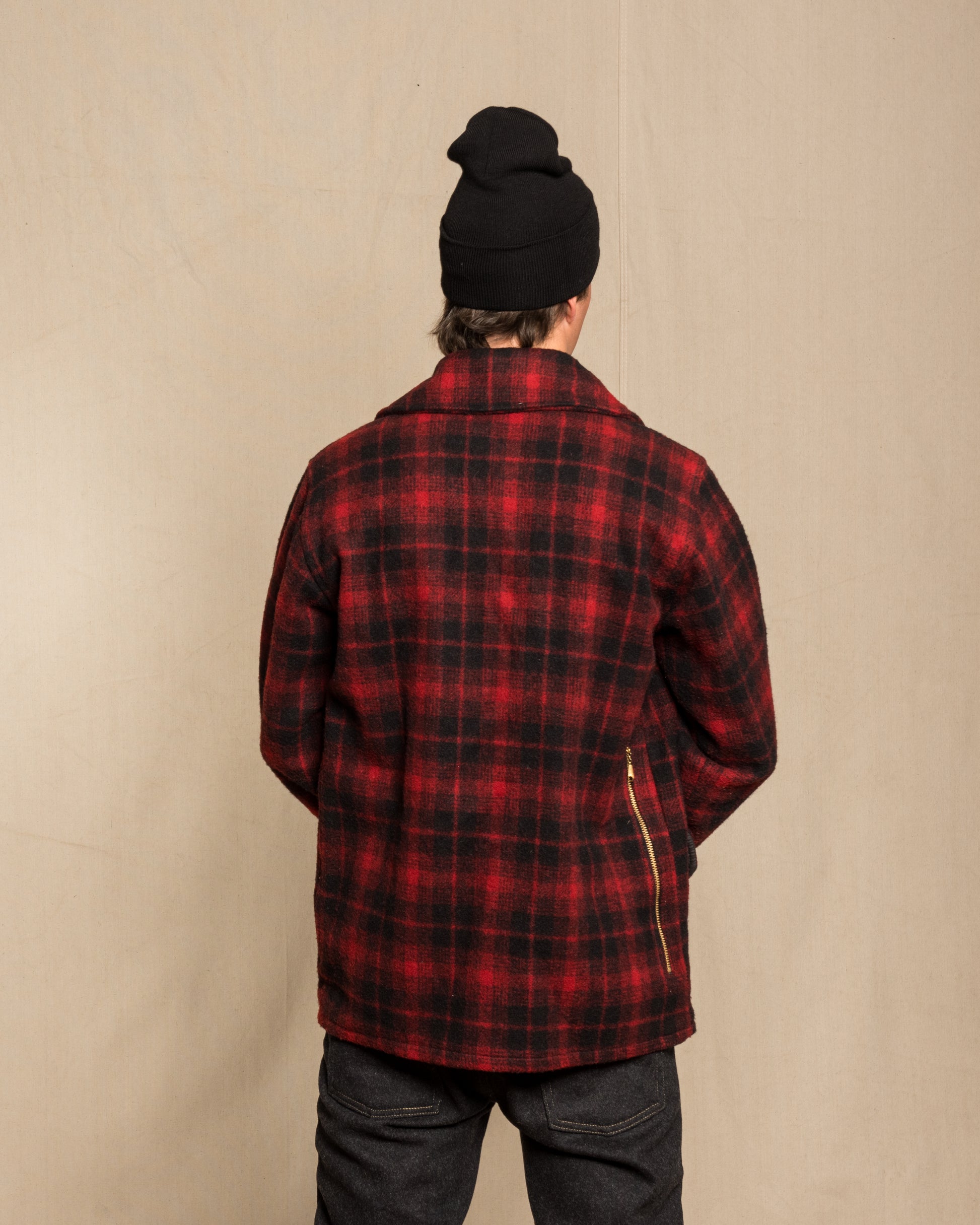 A person viewed from behind wearing a red and black plaid wool flannel jacket with a black knit beanie and dark jeans, standing against a plain beige backdrop.