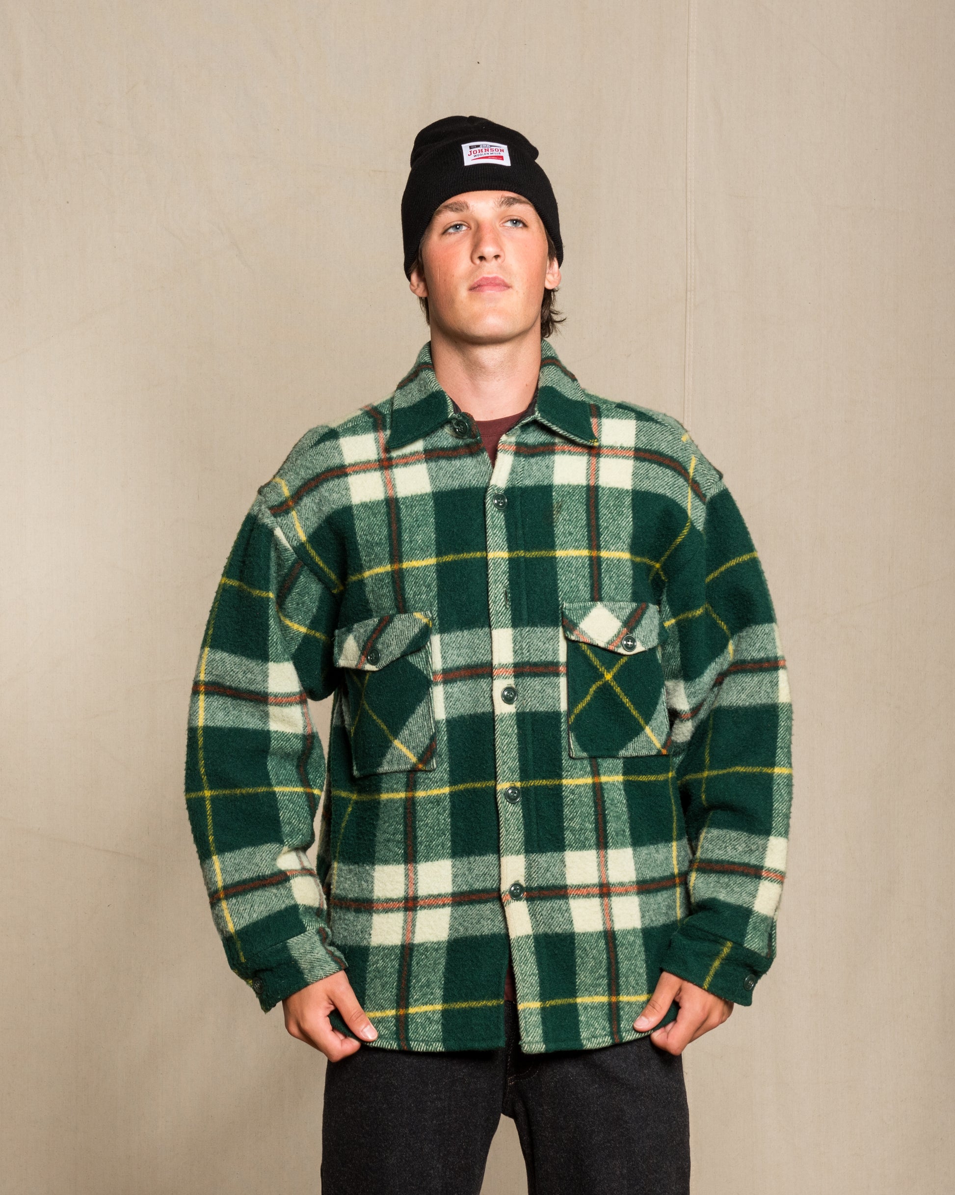 A man wearing a green and white plaid wool button-up shirt jacket with yellow and brown stripes, a black beanie, and dark pants, standing against a light beige wood-paneled background.
