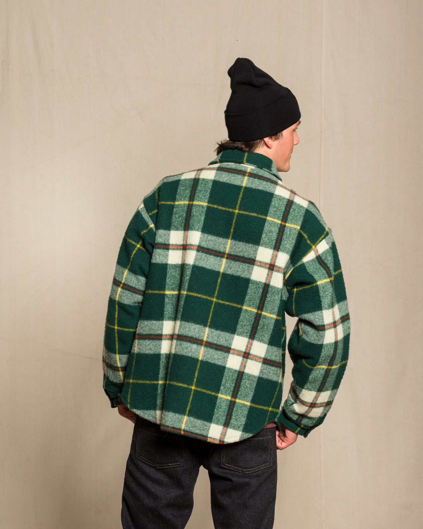 A person shown from the back wearing a green plaid wool flannel shirt with white and yellow lines, paired with a black beanie and dark jeans, standing against a plain beige backdrop.