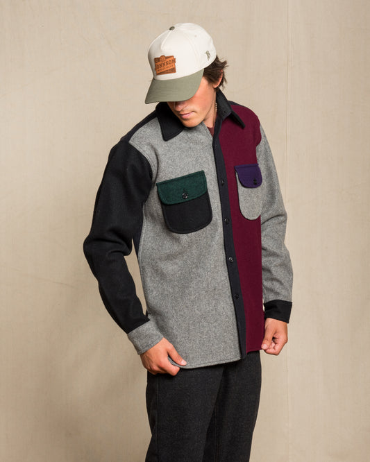 A man wearing a color-blocked button-up shirt with gray, black, burgundy, green, and purple panels, paired with a white and gray baseball cap, standing against a beige textured wall.