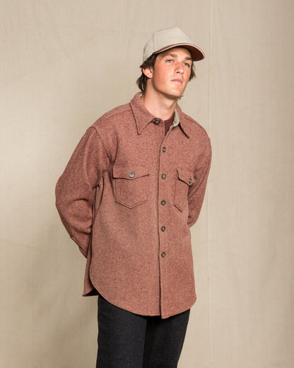 A man wearing a twill wool red button-up shirt jacket with chest pockets, a beige baseball cap, and dark pants, standing against a neutral beige studio backdrop.