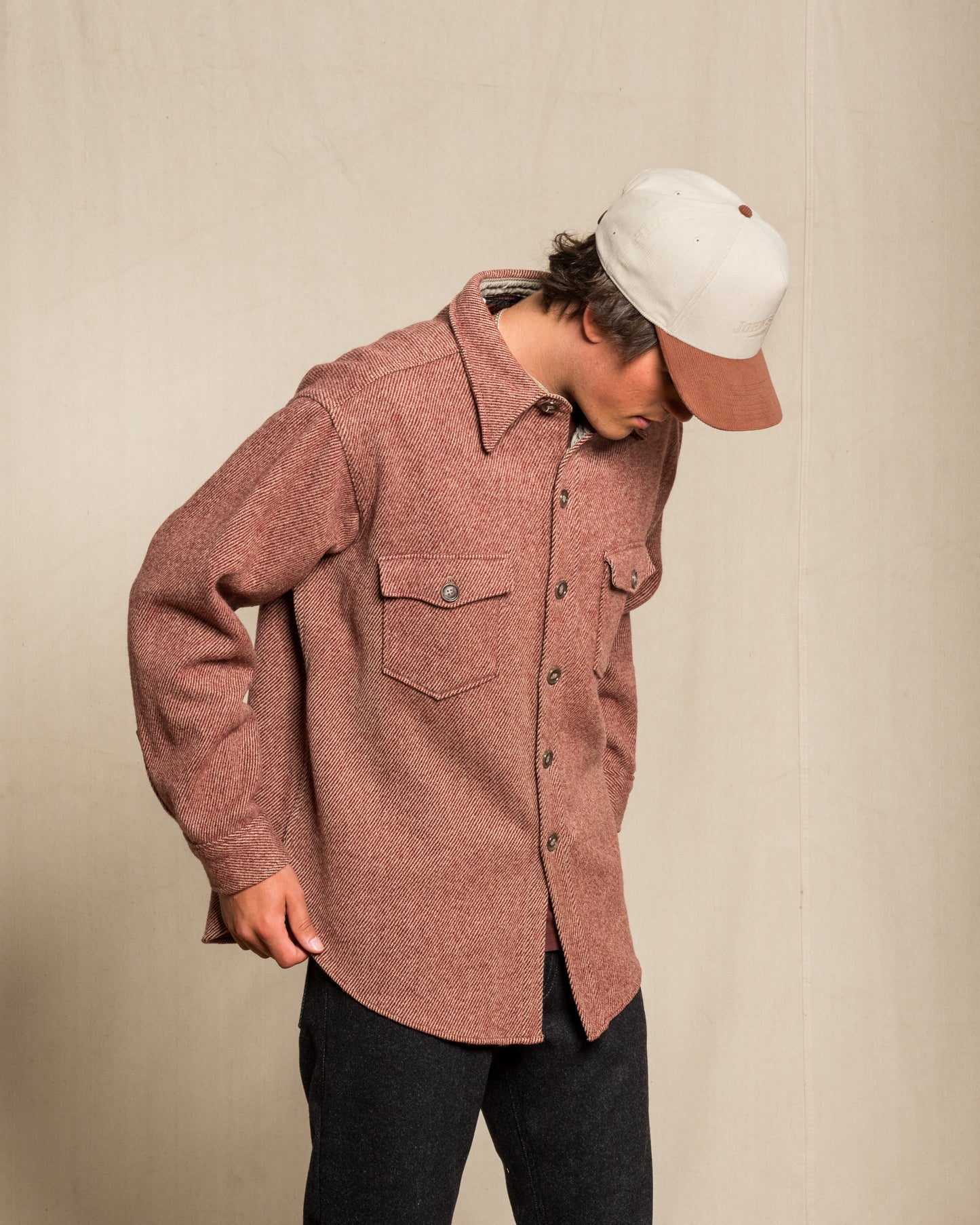 A person wearing a twill wool red button-up shirt, dark jeans, and a white cap with a brown brim, standing against a neutral beige wall.