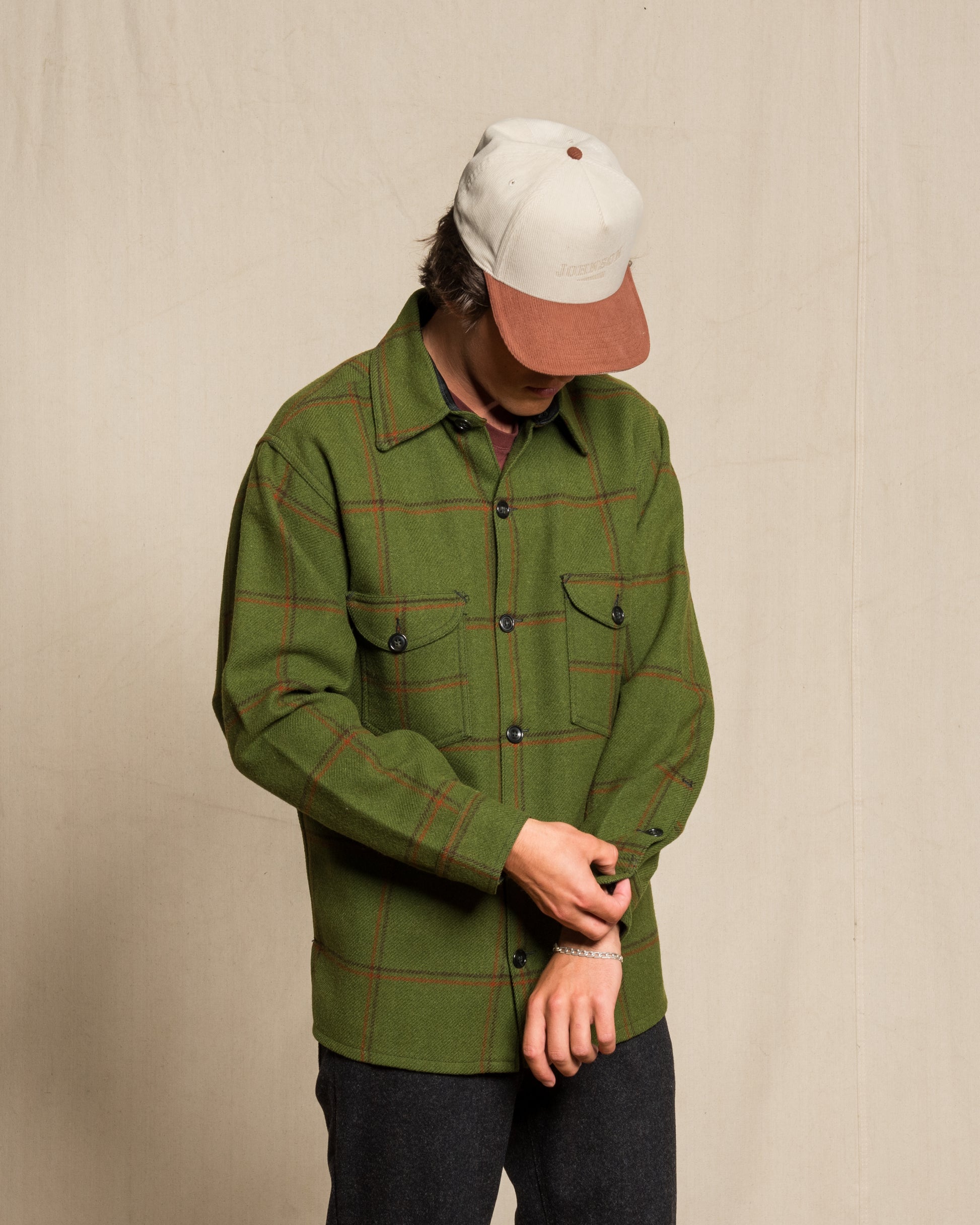 A person wearing an olive green plaid button-up shirt with red lines, a cream and brown baseball cap, and dark pants, standing against a plain beige backdrop.