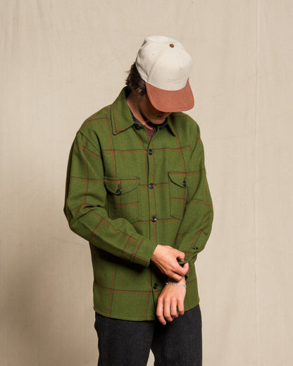 A person wearing an olive green plaid button-up shirt with red lines, a cream and brown baseball cap, and dark pants, standing against a plain beige backdrop.
