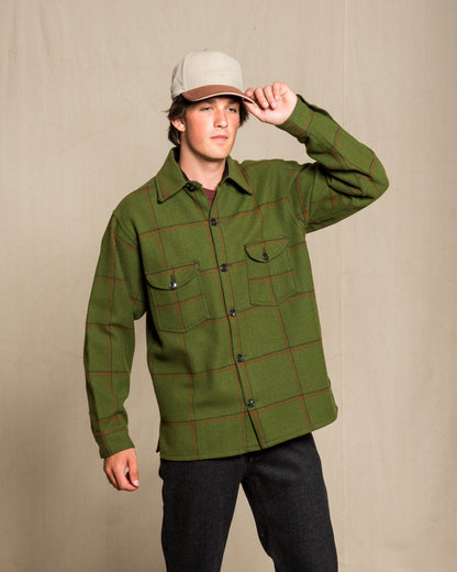 A man wearing an olive green long-sleeve plaid button-up shirt with dark buttons and two chest pockets, paired with dark pants and a beige baseball cap, standing against a neutral beige backdrop.